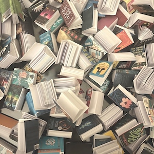 May include: A collection of miniature books, some open, scattered across a dark surface. The books have various colored covers and titles, including "Picture of Dorian Gray" and "Song of Achilles". The pages are white and the overall effect is a literary theme.