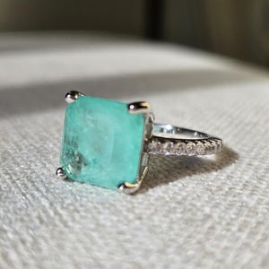 May include: A silver-toned ring featuring a large, square-cut, translucent mint-green gemstone held by four prongs. The band is slender and decorated with a row of small, clear, round-cut stones along the visible side of the metal shank.