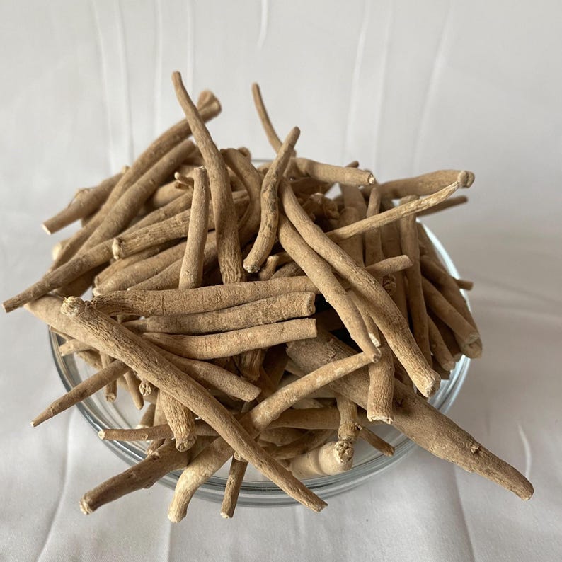 May include: A clear glass bowl filled with dried, beige ashwagandha roots. The roots are long, cylindrical, and have a slightly rough texture. The background is a plain white surface.