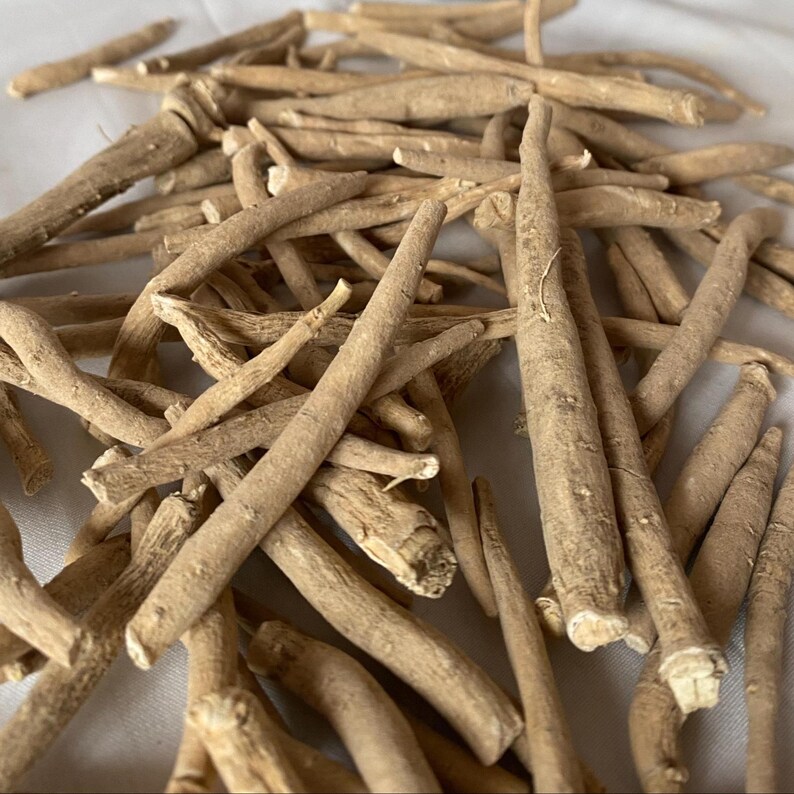 May include: A pile of dried, light brown ashwagandha roots. The roots are various sizes and shapes, with a rough, textured surface. The image is taken from a slightly elevated angle, showing the roots scattered on a white surface.