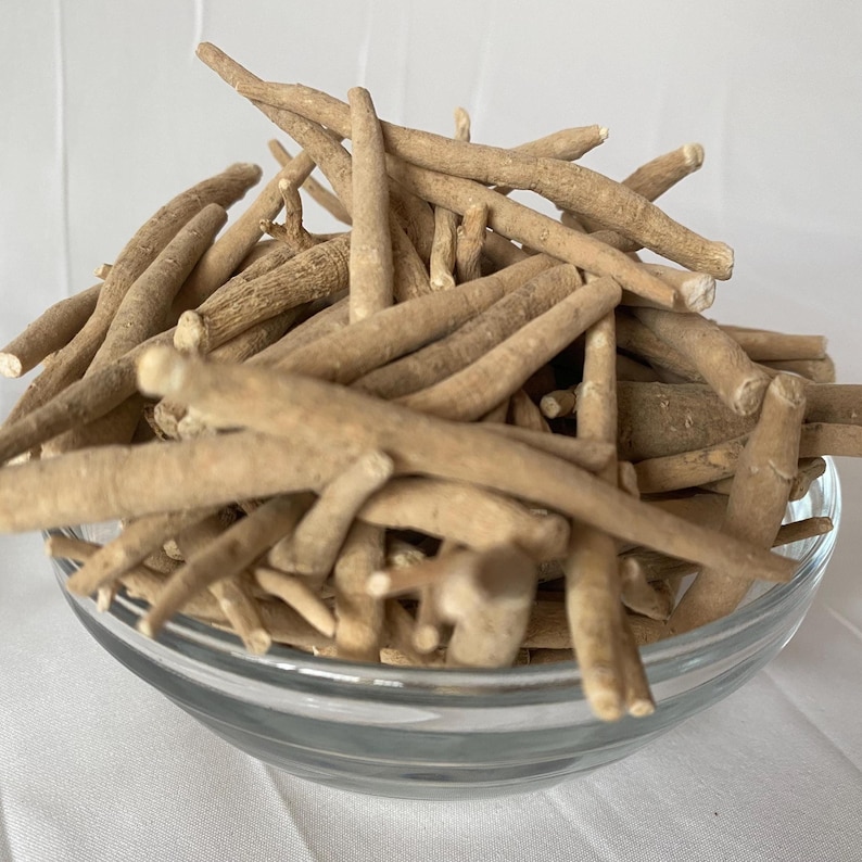 May include: A clear glass bowl filled with light brown ashwagandha roots. The roots are cylindrical and vary in size, with a rough, textured appearance. The bowl sits on a white surface.
