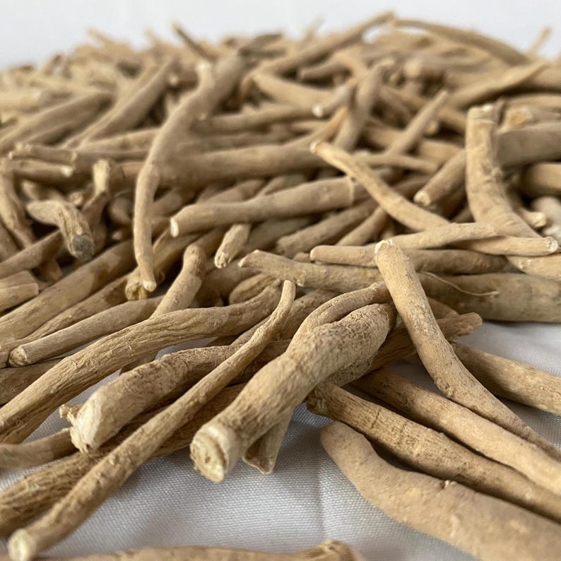 May include: A close-up shot of a pile of dried, beige-colored ashwagandha roots. The roots are long, cylindrical, and have a rough texture. They are arranged in a haphazard manner, with some roots overlapping others. The background is a plain white surface.