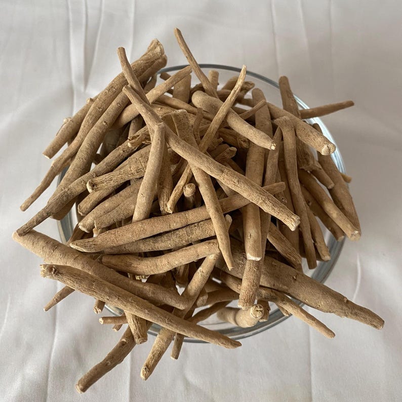 May include: A clear glass bowl filled with numerous light brown, dried ashwagandha roots. The roots are cylindrical, with a rough texture and varying lengths. The bowl sits on a white surface, providing a clean backdrop for the herbal product.