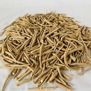 May include: A pile of dried, beige-colored ashwagandha roots. The roots are of varying lengths and thicknesses, with a rough, textured appearance. The image is taken from a slightly elevated angle, showing the roots scattered on a white surface.