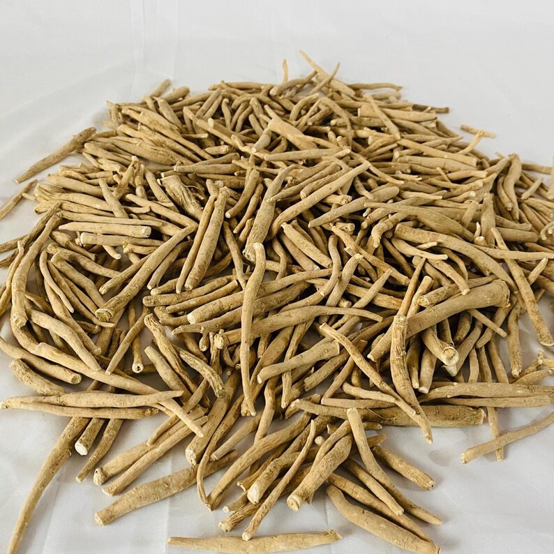 May include: A pile of dried, beige-colored ashwagandha roots. The roots are of varying lengths and thicknesses, with a rough, textured appearance. The image is taken from a slightly elevated angle, showing the roots scattered on a white surface.