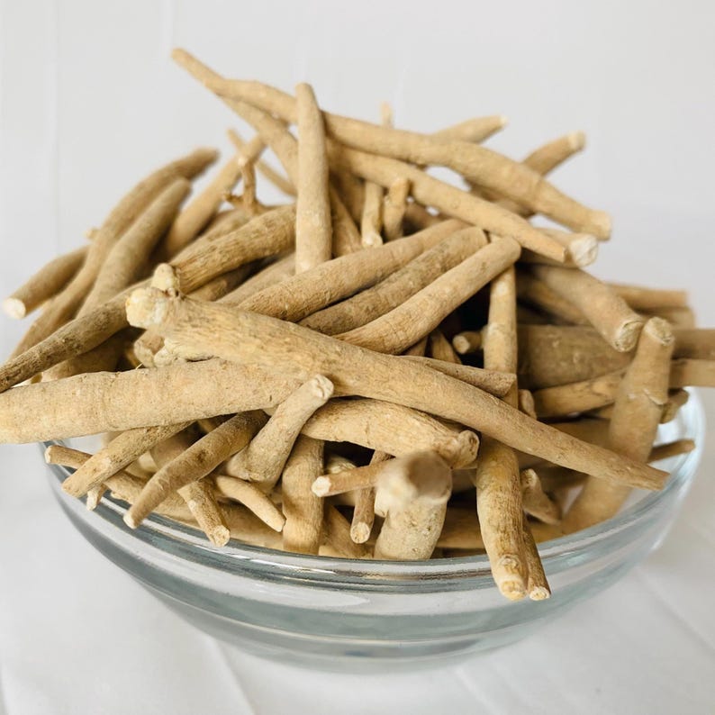 May include: A clear glass bowl filled with tan-colored, dried ashwagandha roots. The roots are cylindrical, with a rough texture and varying lengths. The bowl sits on a white surface, and the background is a soft white.