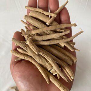 May include: A handful of dried, light brown ashwagandha roots. The roots are long, cylindrical, and have a rough texture. The image is taken from a slightly elevated angle, showing the roots in detail. The background is a plain white surface.