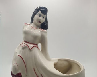 Puede incluir: Un macetero de cerámica blanca con forma de mujer con un vestido blanco y detalles rojos. La mujer tiene el pelo negro y un lazo rojo en el vestido.
