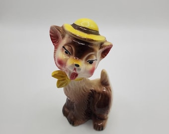 May include: A vintage ceramic figurine of a dog with a yellow hat and bow tie. The dog has brown and tan fur, pink cheeks, and a playful expression. The figurine is standing upright.