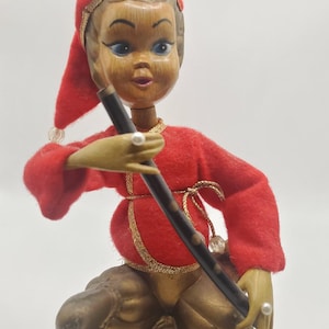 May include: A vintage figurine of a seated figure in a red outfit with gold trim, holding a black flute. The figure has a wooden face, blue eyes, and a red hat. The base is brown and gold.