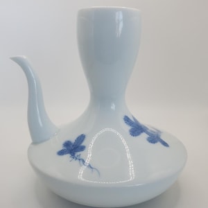 Puede incluir: Una botella de sake de cerámica blanca con un diseño floral azul. La botella tiene un pico y una base ancha.