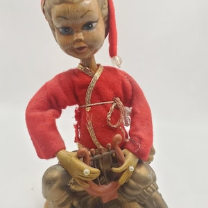May include: A vintage figurine of a seated figure with a wooden head and hands, wearing a red outfit with gold trim and a red cap. The figure is holding a small harp-like instrument. The base is a mottled brown color.