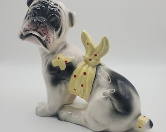 Peut inclure: Une figurine en céramique d'un bouledogue noir et blanc portant un bandage jaune et rouge à pois. Le bouledogue est assis, la tête inclinée sur le côté et la bouche légèrement ouverte.