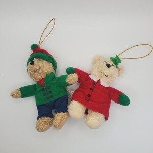 Könnte beinhalten: Zwei Plüsch-Bären-Weihnachtsbaumschmuck. Ein Bär trägt eine grüne Jacke, blaue Hose und einen rot-grünen Hut. Der andere hat einen roten Mantel mit grünen Akzenten und einen Stechpalmenzweig. Beide haben goldene Schnuraufhänger.