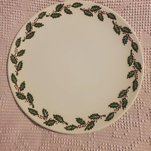 May include: Round white ceramic plate featuring a border of green leaves and red berries. The plate's design is simple and elegant, suitable for serving food or as a decorative item. The plate is circular.