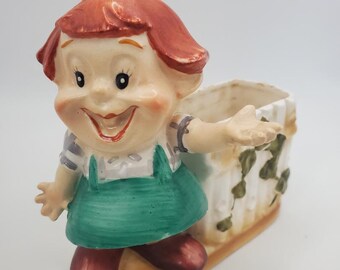 May include: A ceramic planter shaped like a girl with red hair, wearing a green dress and brown shoes. The planter has a white picket fence design and is holding out her hand.