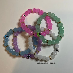 May include: A set of five colorful beaded bracelets with gold accents and evil eye charms. The bracelets are in shades of pink, green, blue, purple, and white. "BUNDLE INCLUDES ALL 5 BRACELETS." is written below the bracelets.