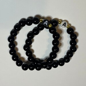 May include: Two black beaded bracelets with gold accents and white letter beads spelling "A".