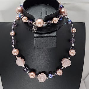 Pode incluir: Um conjunto delicado de colar e pulseira com pérolas rosa claro, contas em forma de rosa e cristais roxos iridescentes. O colar e a pulseira são enfiados em um fio de prata.