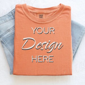 May include: A folded orange t-shirt with the text "YOUR Design HERE" on the front. The t-shirt is on top of a pair of blue jeans.