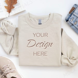 May include: A beige sweatshirt with the text "YOUR Design HERE" printed on the front. The sweatshirt is laid flat on a white surface with a pair of beige sneakers, a book, and a pair of blue jeans.