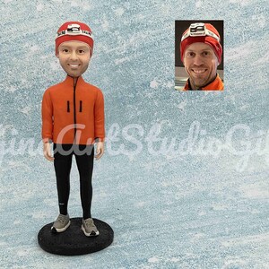 May include: A custom bobblehead figurine of a person wearing a red jacket, black pants, and a red and white hat with a headlamp. The figurine is standing on a black base.