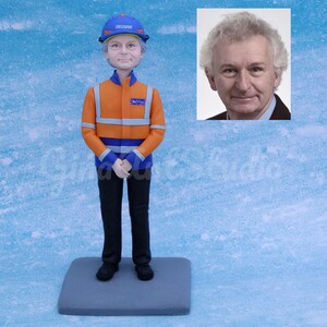 May include: A figurine of a person in an orange and blue safety uniform, blue hard hat, and black pants. The figurine stands on a gray base. A photo of a man in a suit is in the upper right corner. The background is a light blue.