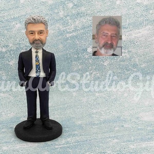 May include: A custom-made figurine of a person wearing a dark blue suit with a patterned tie. The figurine is standing on a black base.