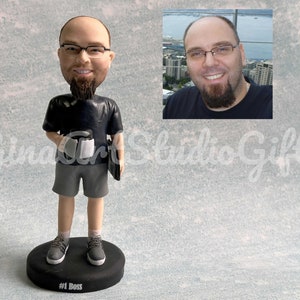 May include: A custom figurine of a person with a beard and glasses, wearing a black t-shirt, gray shorts, and sneakers. The figurine is holding a coffee cup and a laptop. The base of the figurine is black and has the text "#1 Boss" written on it.