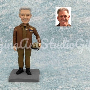 Puede incluir: Una figura personalizada de una persona que lleva una chaqueta marrón, pantalones verdes y un sombrero de estilo militar. La figura sostiene un casco verde en su mano derecha. La figura está de pie sobre una base gris.
