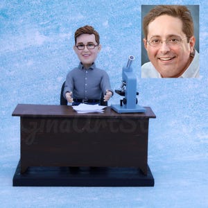 Può includere: Una statuina in miniatura di un uomo con camicia grigia e occhiali seduto a una scrivania marrone. Un microscopio blu e dei fogli sono sulla scrivania. Una piccola foto di un uomo con gli occhiali è nell'angolo in alto a destra. La scrivania ha il testo "ginaArtSt".