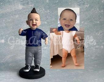 May include: A custom figurine of a baby with a mohawk hairstyle wearing a blue shirt, gray pants, and black sneakers. The figurine is standing on a black base. The baby's face is smiling.