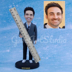 May include: A custom bobblehead figurine of a man in a gray suit holding a 20-inch ruler. The figurine is on a black base with the name "DIVORO" and a small photo of the man. The background is a light blue textured surface.