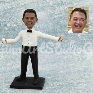 May include: A figurine of a person wearing a white shirt and black trousers, holding a small white object in one hand and a stack of wooden sticks in the other. The figurine is standing on a black base. A smaller image of the person's face is in the background.