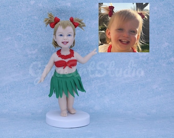 Puede incluir: Una figura personalizada de una niña pequeña con un atuendo de hula, con un top y collar rojos, una falda verde y cabello rubio con lazos rojos. Una pequeña foto de la niña está en la esquina superior derecha. La figura está sobre una base blanca.