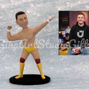 May include: A custom figurine of a wrestler in a yellow and red wrestling outfit, striking a pose with one arm raised and the other clenched in a fist. The figurine is standing on a black base. The background shows a television screen with a person sitting on a couch.