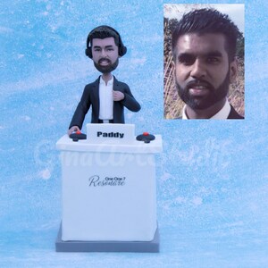 May include: A white figurine of a DJ wearing headphones and a black suit, standing behind a white console. The console has the name "Paddy" on it and the text "One One 7 Resonate". A photo of a man is in the upper right corner.