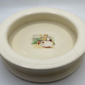 May include: A white ceramic dish with a raised rim. The dish features a small illustration of a girl in a dress holding a tray of food. The text "The Nurse" is printed below the illustration.