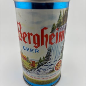 May include: A blue and silver aluminium can of Bergheim Beer featuring a picture of a waterfall and a red horse-drawn carriage. The text on the can reads "Bergheim Beer" and "Brewed with pure mountain spring water from the Penna. Dutch Country."