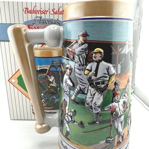 May include: A ceramic beer stein with a baseball bat-shaped handle. The stein features a baseball scene with players in vintage uniforms. The stein has the text "Budweiser Salutes America's Pastime".