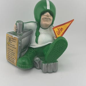 May include: A ceramic figurine of a football player wearing a green helmet and uniform, seated with a television set. The figurine holds a pennant that says "Win." The base of the figurine reads "Armchair Quarterback." The television set has text that reads "Please deposit a quarter."