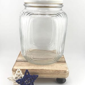May include: A clear glass jar with a white lid and a gold-colored band. The jar sits on a small, light-colored wooden platform with two decorative star-shaped objects in white and navy blue.