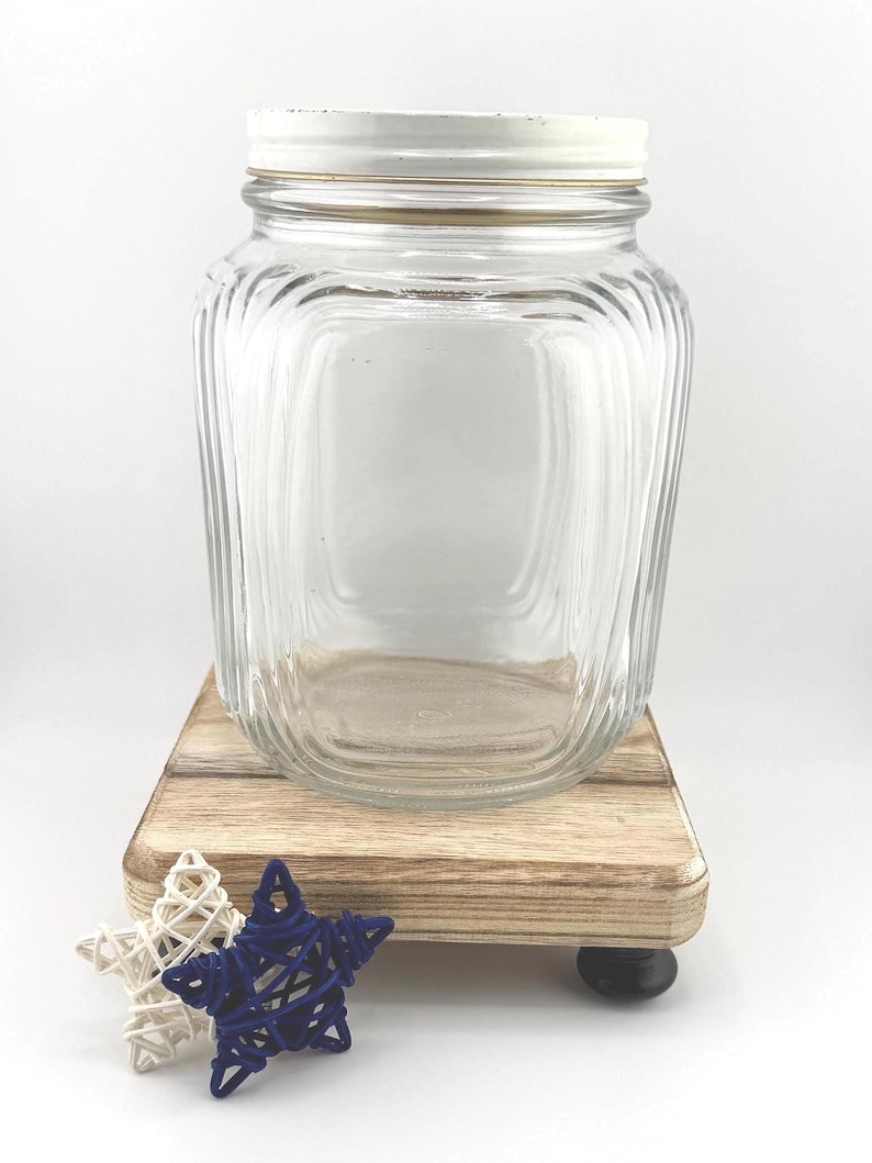 May include: A clear glass jar with a white lid and a gold-colored band. The jar sits on a small, light-colored wooden platform with two decorative star-shaped objects in white and navy blue.