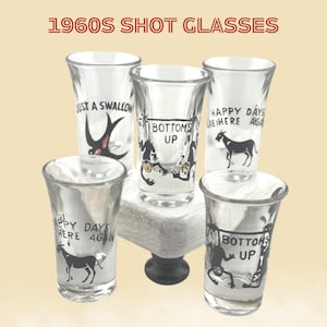 May include: Five vintage shot glasses with black and white graphics and text. The text includes "Just a Swallow", "Bottoms Up", and "Happy Days Here Again". The glasses are clear and sit on a small, white pedestal. The words "1960s Shot Glasses" are at the top.