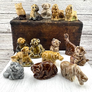 Pode incluir: Uma coleção de estatuetas de animais de cerâmica em várias poses. As estatuetas incluem uma coruja, cães, um coala, macacos, um urso, um coelho, um gato, um hipopótamo, um guaxinim e um elefante. As estatuetas são em tons de castanho, bege e cinzento.