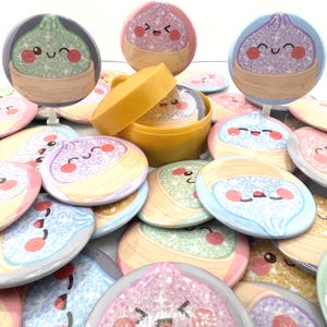 May include: A collection of circular pin-back buttons featuring glittery, smiling dumpling characters in bamboo steamers, alongside a small yellow plastic container holding one of the buttons. The designs include green, purple, and pink dumplings.
