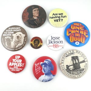 May include: A collection of vintage pinback buttons with various designs and text. Buttons include images of a polar bear, a portrait, and text such as "Are we having fun YET?" and "Support Your Right to Arm Bears."
