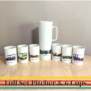 Puede incluir: Un jarro de cerámica blanca y seis tazas a juego. Cada taza y el jarro están decorados con ilustraciones de coches antiguos en varios colores. El juego se exhibe sobre una superficie de madera.