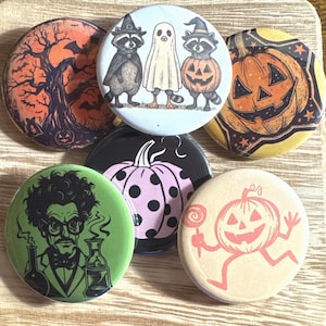 May include: A collection of Halloween-themed pinback buttons. Designs include a spooky tree, jack-o'-lanterns, raccoons in witch hats, a ghost, a mad scientist, and a pink polka-dotted pumpkin. The buttons are round and colorful.