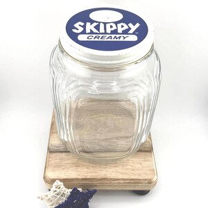 May include: A clear glass jar with a white and blue lid that reads "Skippy Creamy." The jar has a square base and a ridged design. It sits on a small wooden stand with decorative accents.